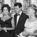 Eddie Fisher, wearing a tuxedo, stands with arm around his wife, Debbie Reynolds and smiles while looking at Elizabeth Taylor, Las Vegas, Nevada, 1958. The next year Fisher left Reynolds and married Taylor