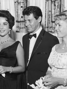 Eddie Fisher, wearing a tuxedo, stands with arm around his wife, Debbie Reynolds and smiles while looking at Elizabeth Taylor, Las Vegas, Nevada, 1958. The next year Fisher left Reynolds and married Taylor